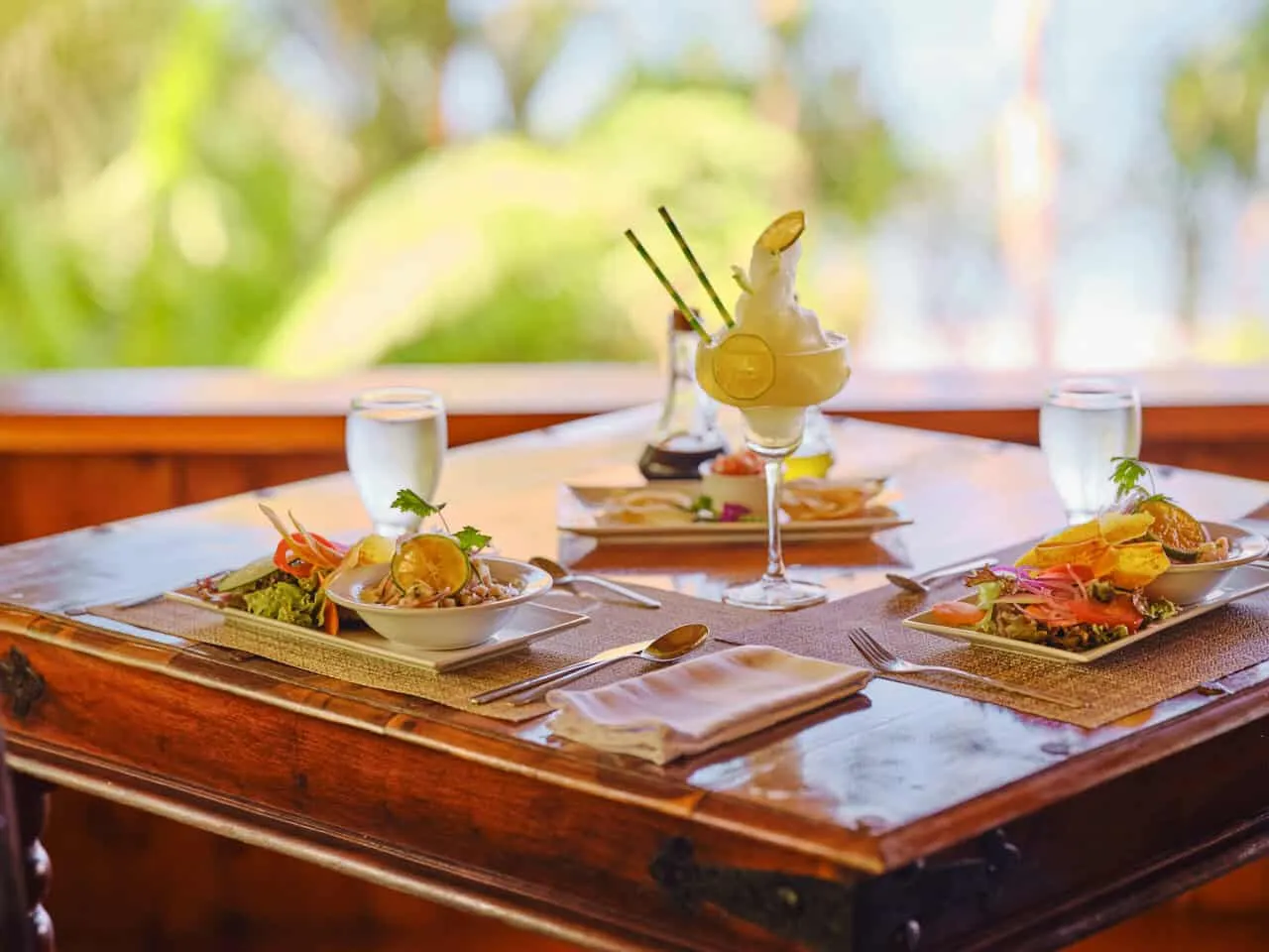 A wooden table set with plates of salad, glasses of water, a dessert in a tall glass, and cutlery, with greenery visible in the blurred background.