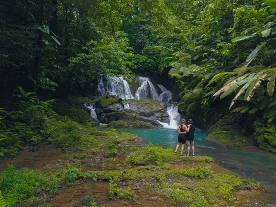 Two people standing in front of a waterfall in the jungle.