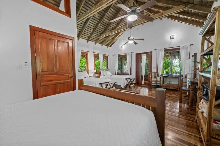 A spacious bedroom with two white-covered beds, wooden furniture, ceiling fans, and large windows providing views of green foliage outside.
