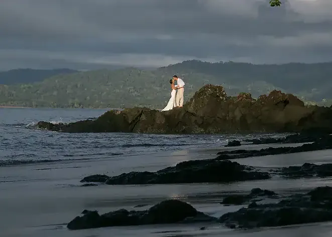 A bride and groom standing on a rock in front of the ocean.