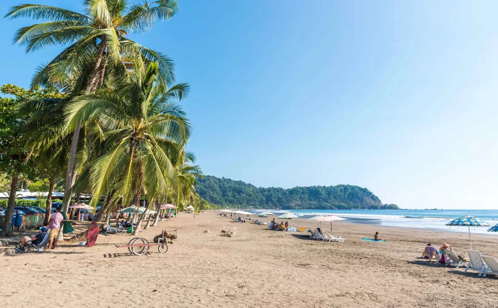 Tropical Jaco Beach scene in Costa Rica with palm trees, lounging visitors, and clear blue skies.