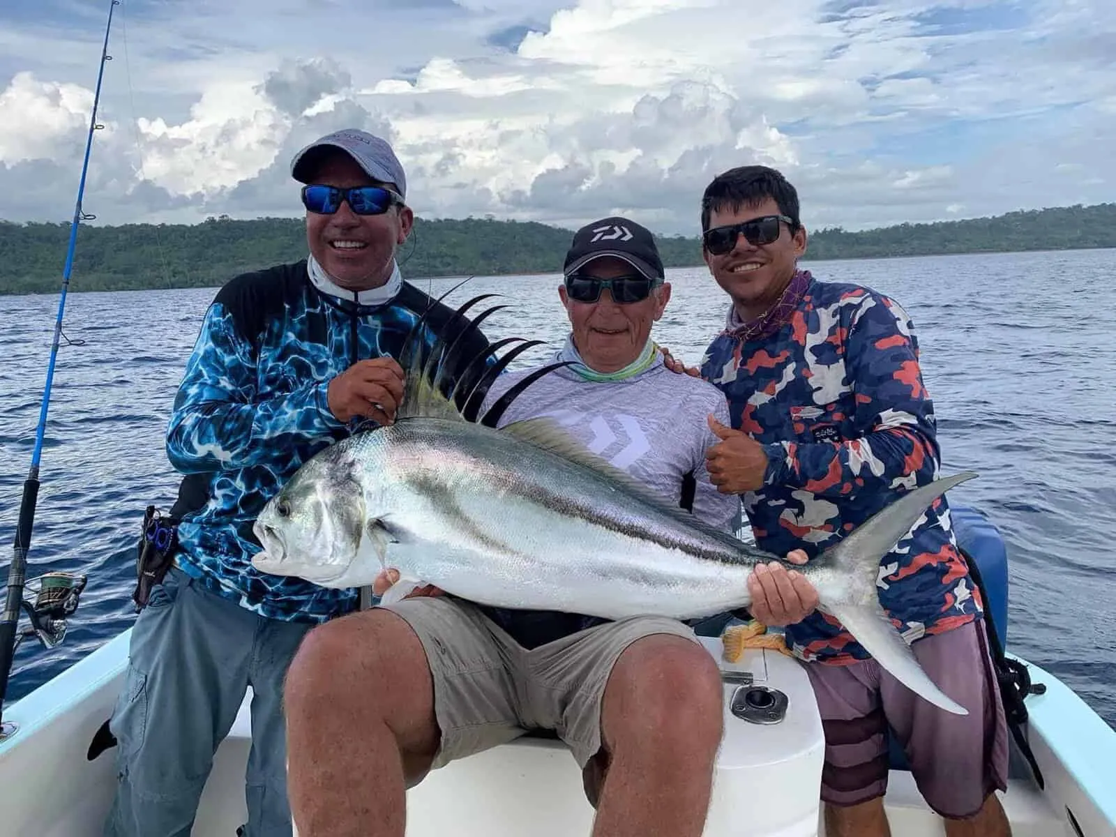Three men on a boat holding a large fish, smiling, with a cloudy sky and calm water in the background.
