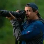 A man using a dslr camera to take photos outdoors, standing in a lush green setting.