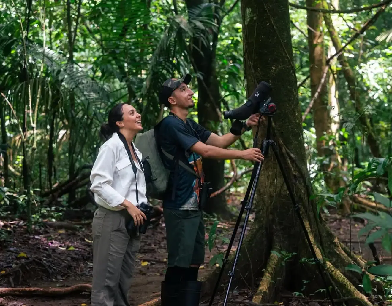 Two people in a rainforest look up while one adjusts a camera with a telephoto lens on a tripod, surrounded by dense green foliage during Guided Tours & Activities in Drake Bay, Costa Rica.