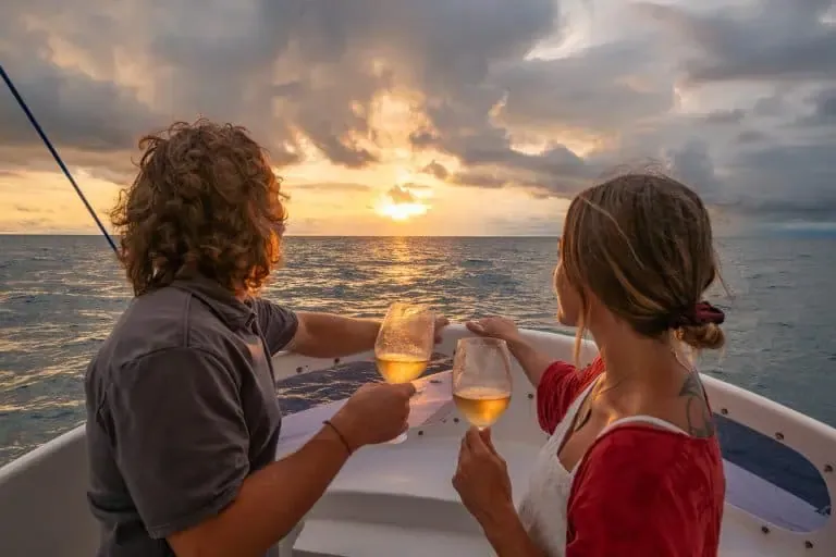 Two people holding wine glasses watch the sunset over the ocean from the deck of a boat, with dramatic clouds in the sky.