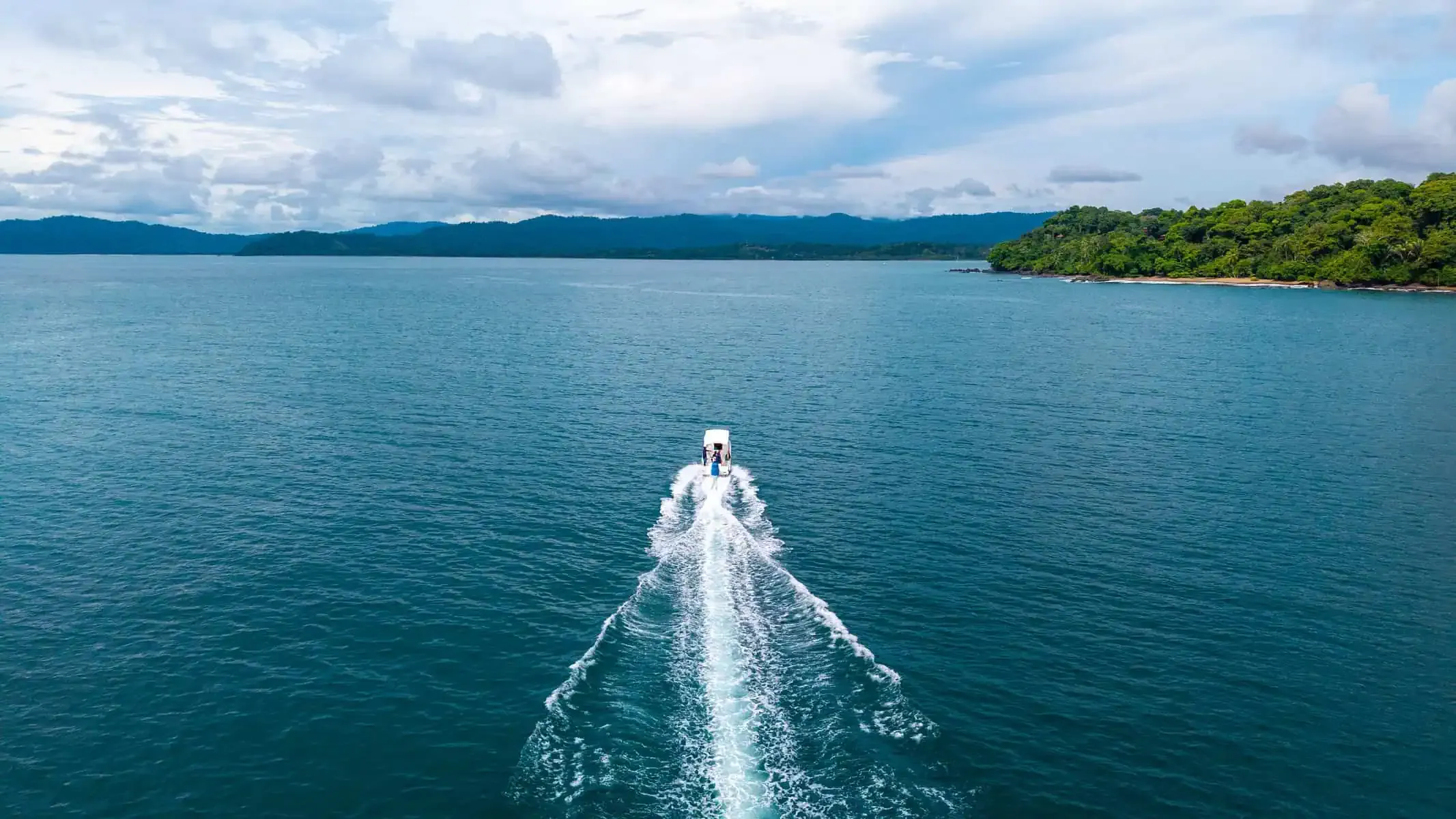 A small motorboat moves through calm blue water, leaving a white wake behind it, with a forested shoreline and mountains visible in the distance under a cloudy sky.