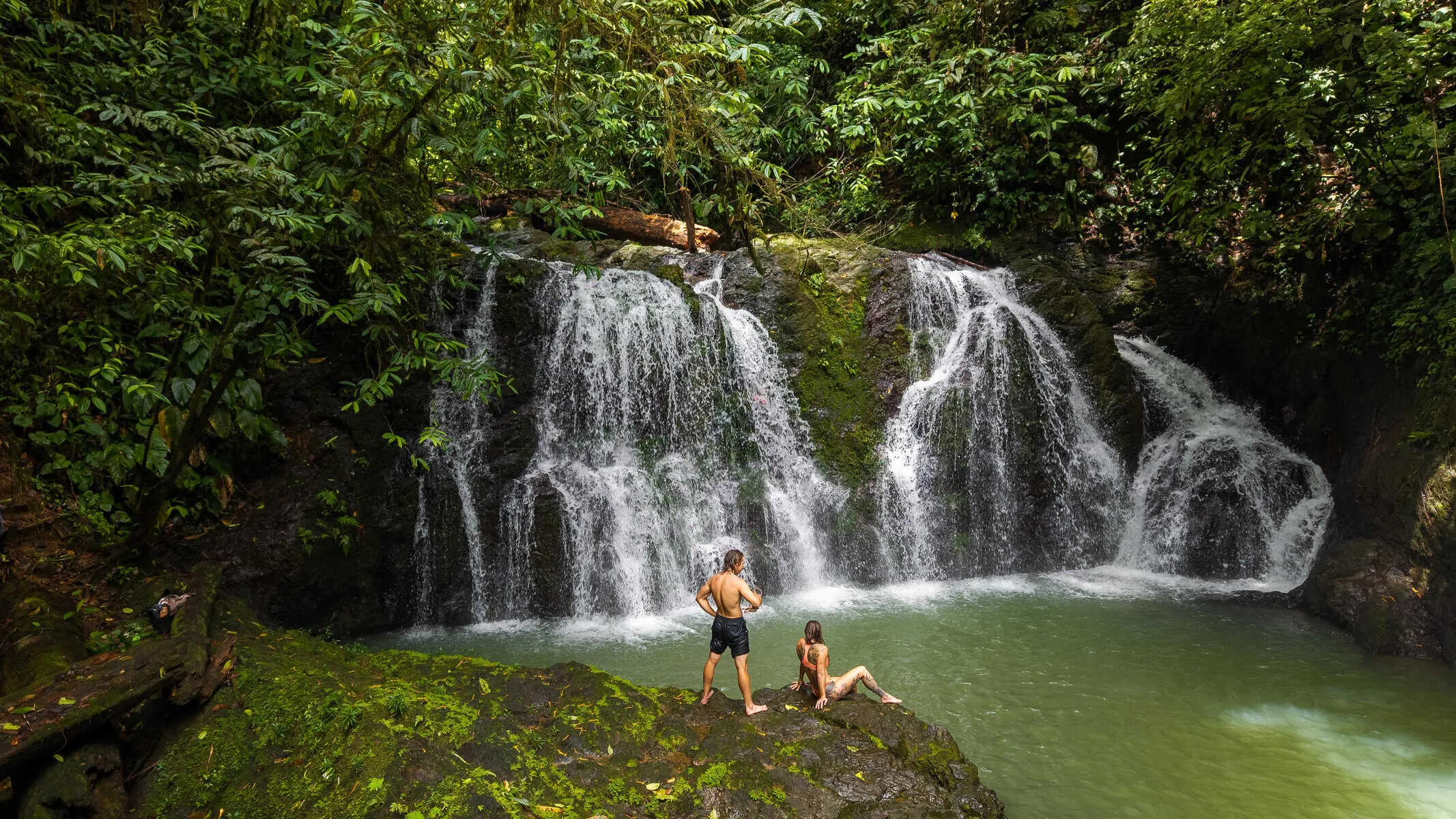 Two people stand on rocks by a small jungle waterfall flowing into a green pool, surrounded by dense, leafy vegetation.