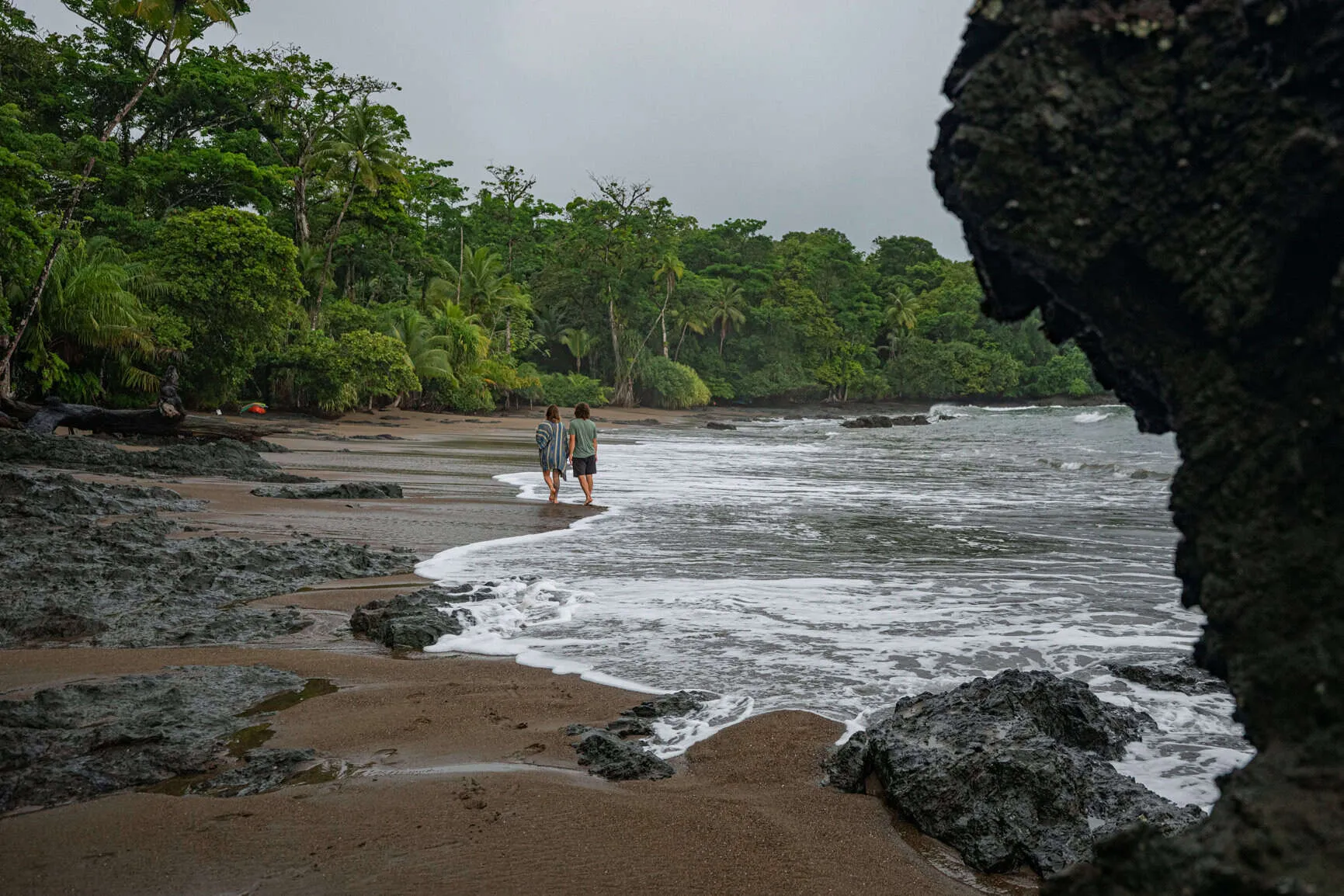 Two people walk along a rocky, tree-lined beach with gentle waves under an overcast sky.