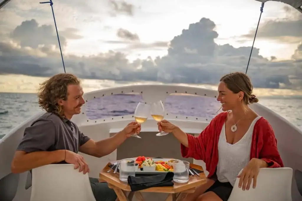 Two people sit at a small table on a boat at sunset, clinking wine glasses over plates of food with the ocean and clouds in the background.