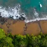 Aerial view of a tropical beach with turquoise waves, rocky shoreline, golden sand, dense green trees, and a white boat in the water.