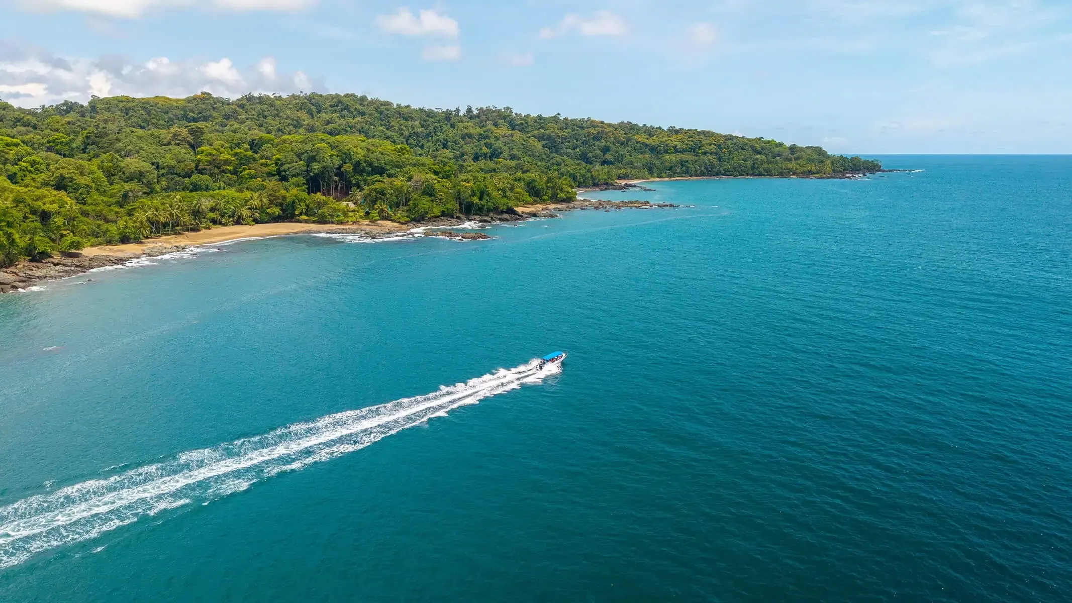 A speedboat moves across turquoise water near a forested coastline with a sandy beach and rocky shoreline under a partly cloudy sky.