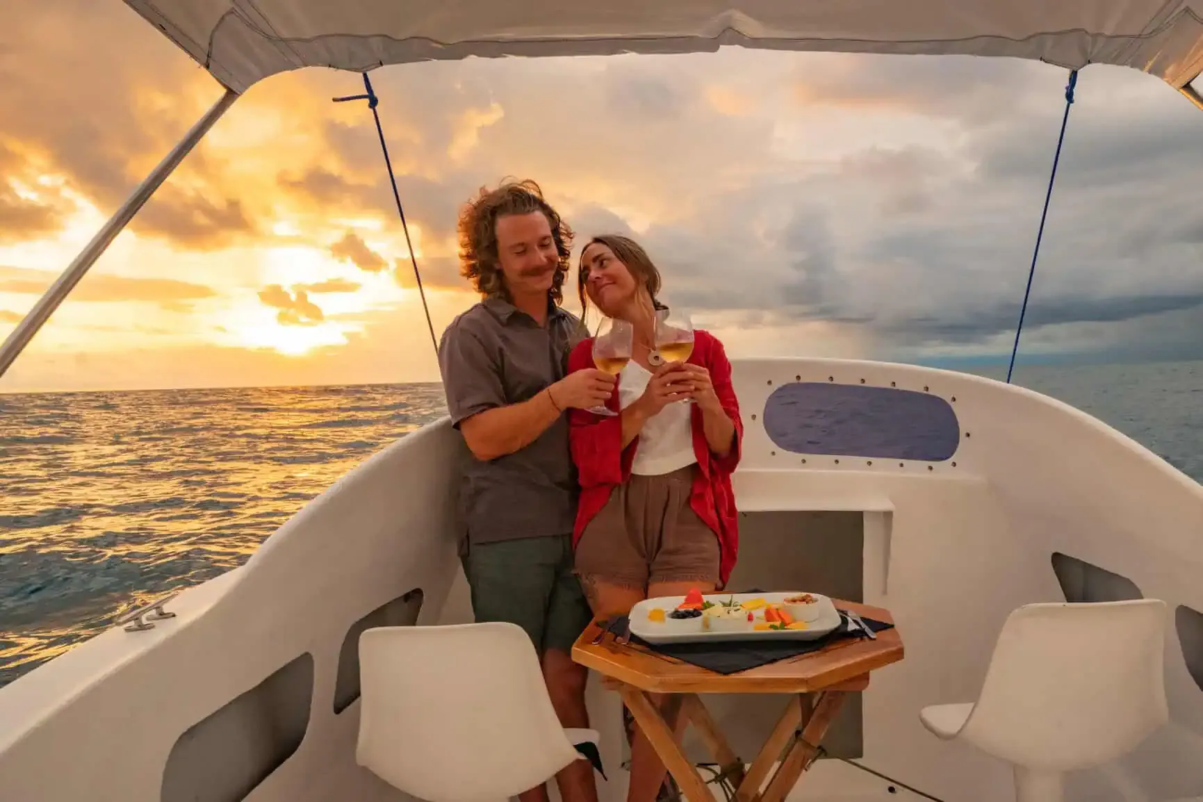 Two people stand on a boat at sunset, clinking glasses and smiling, with a tray of food on a small table between them and the ocean in the background.