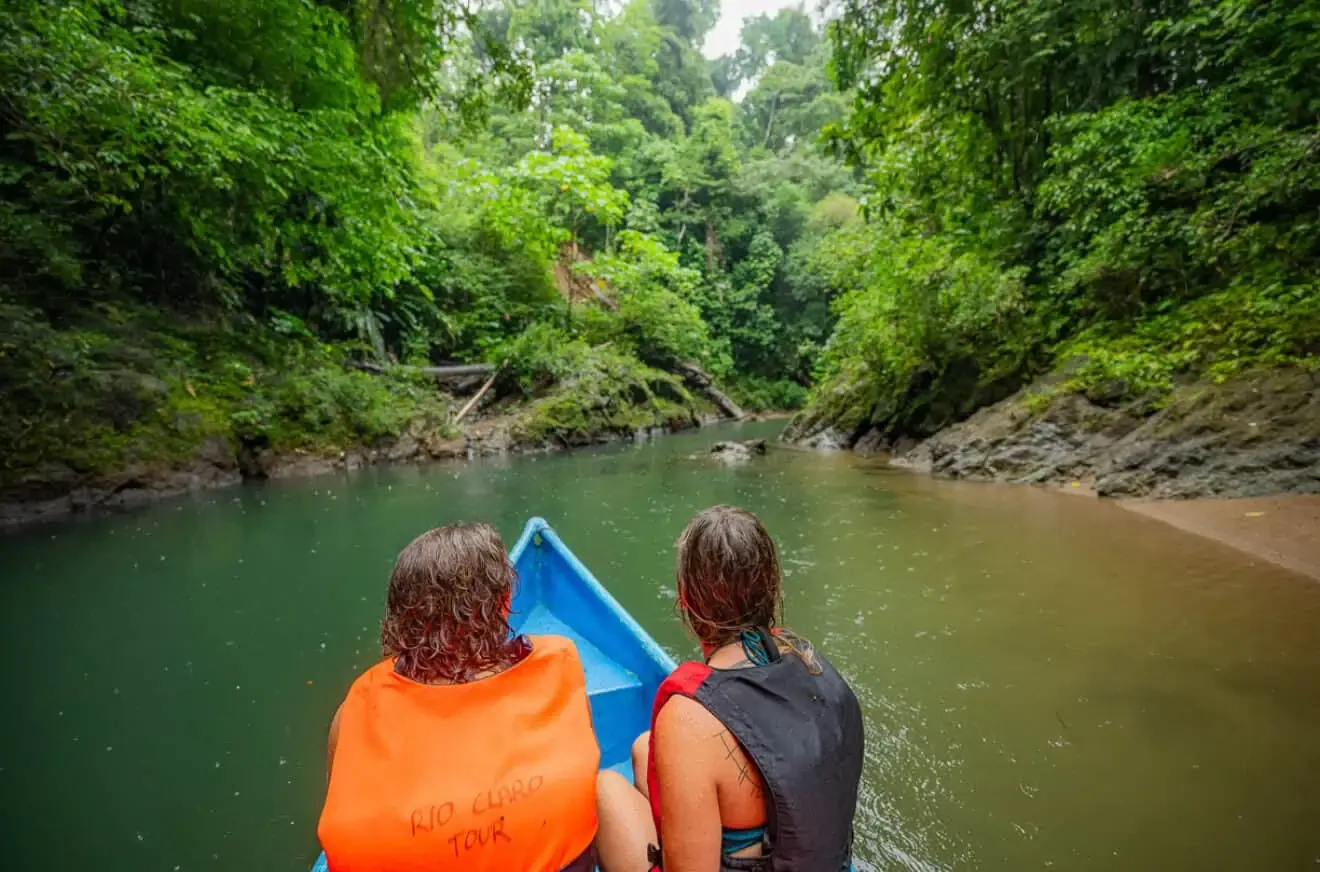 Two people wearing life jackets sit at the front of a blue boat, traveling down a lush, green river surrounded by dense forest.