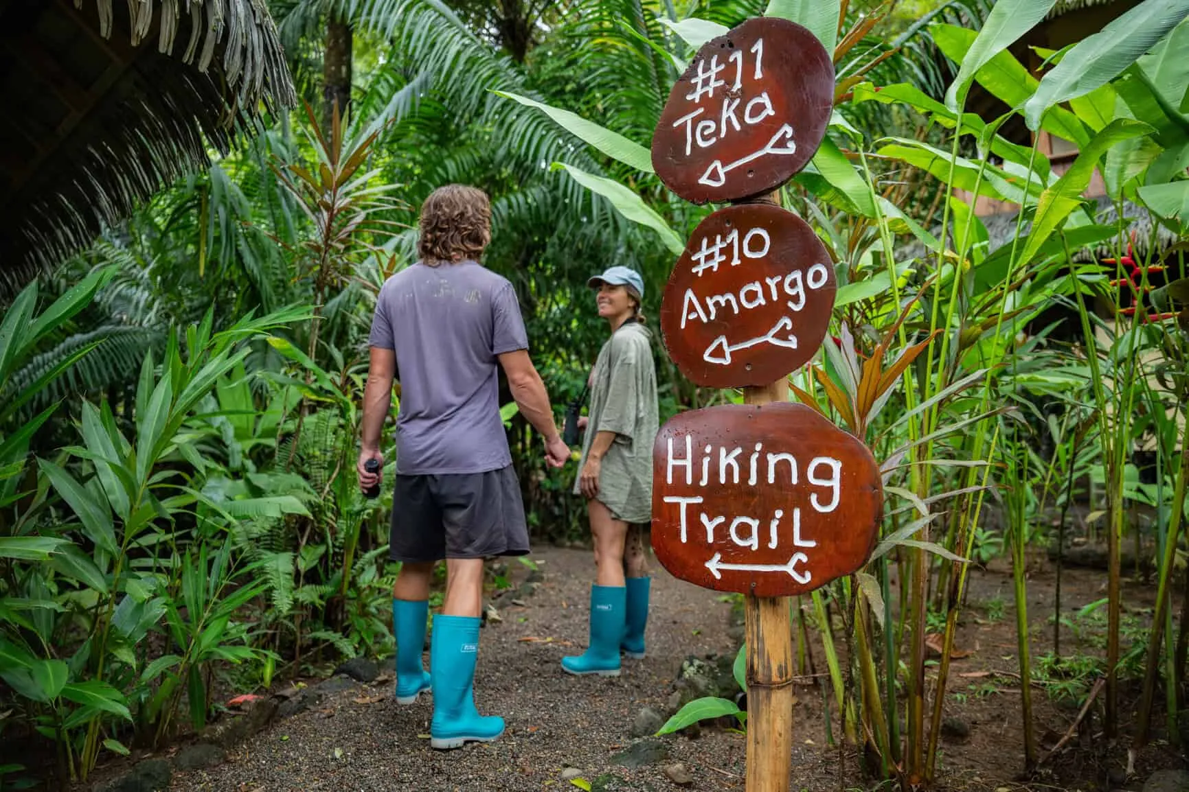 Two people in blue boots stand on a jungle path near wooden signs pointing to cabins #11, #10, and a hiking trail, surrounded by lush tropical vegetation.