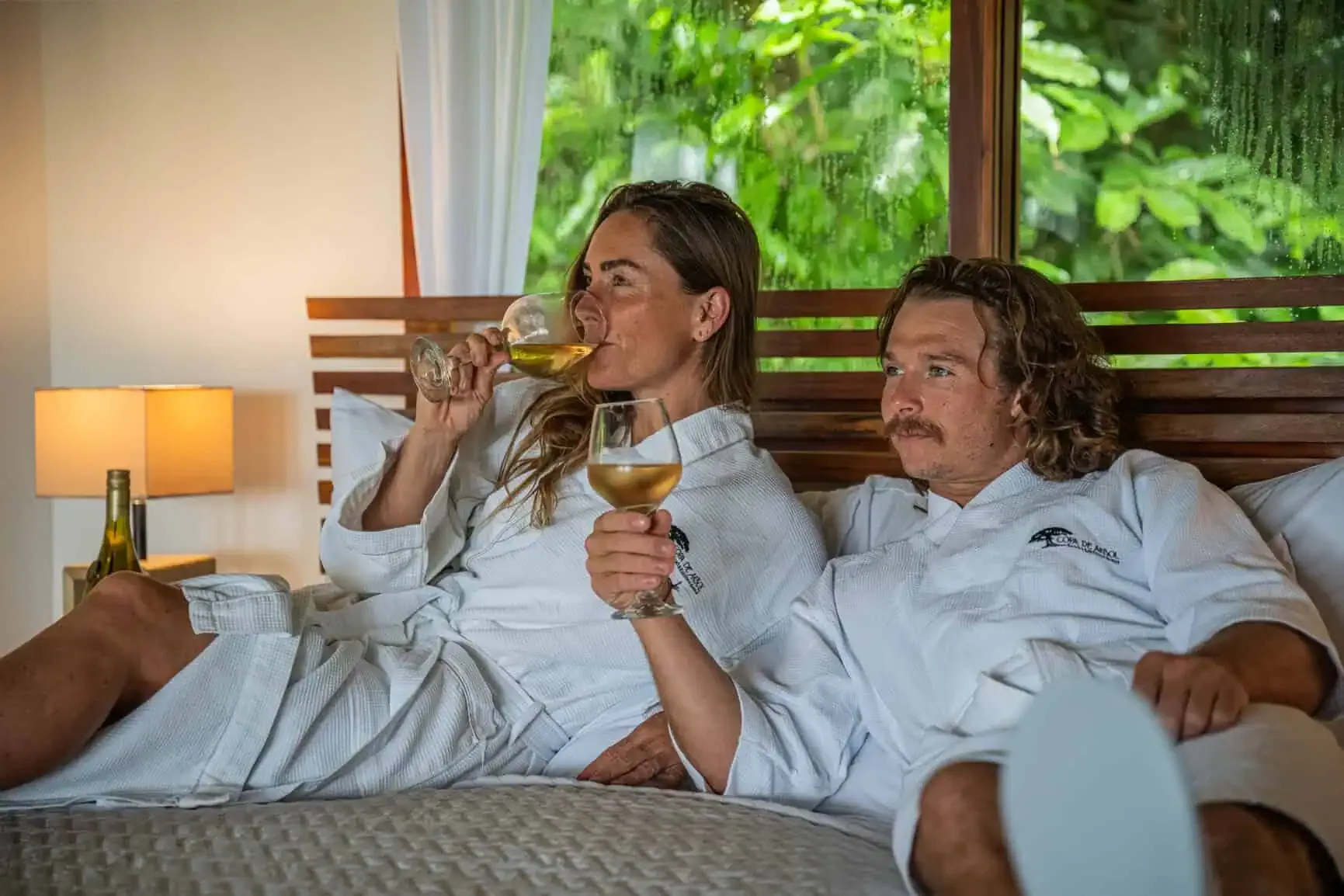 Two people in white robes relax on a bed, each holding a glass of white wine, with a bottle on the nightstand and greenery visible through the window.