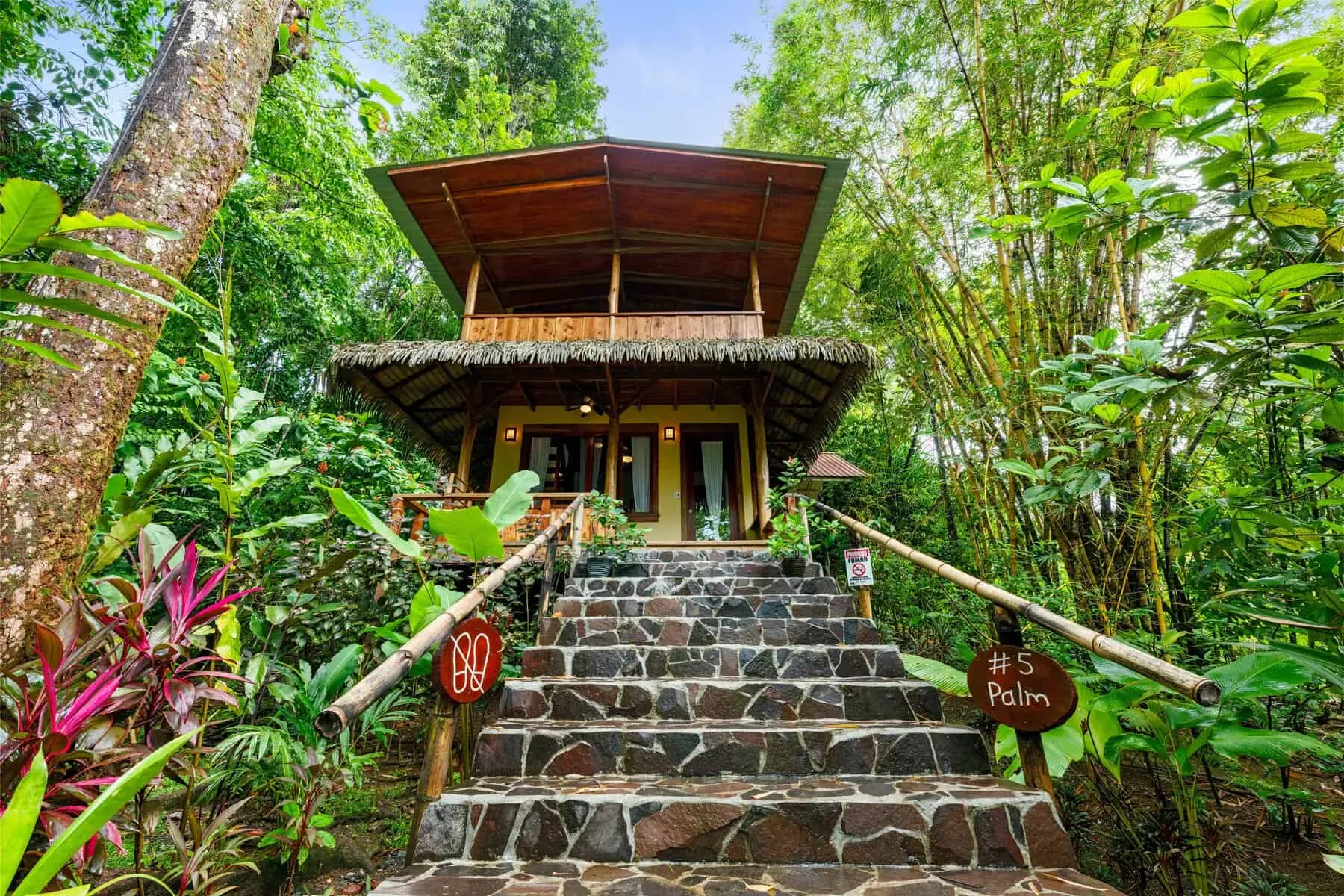 A two-story wooden cabin with a thatched roof sits among lush tropical plants, stone steps leading to the entrance, and a sign reading 