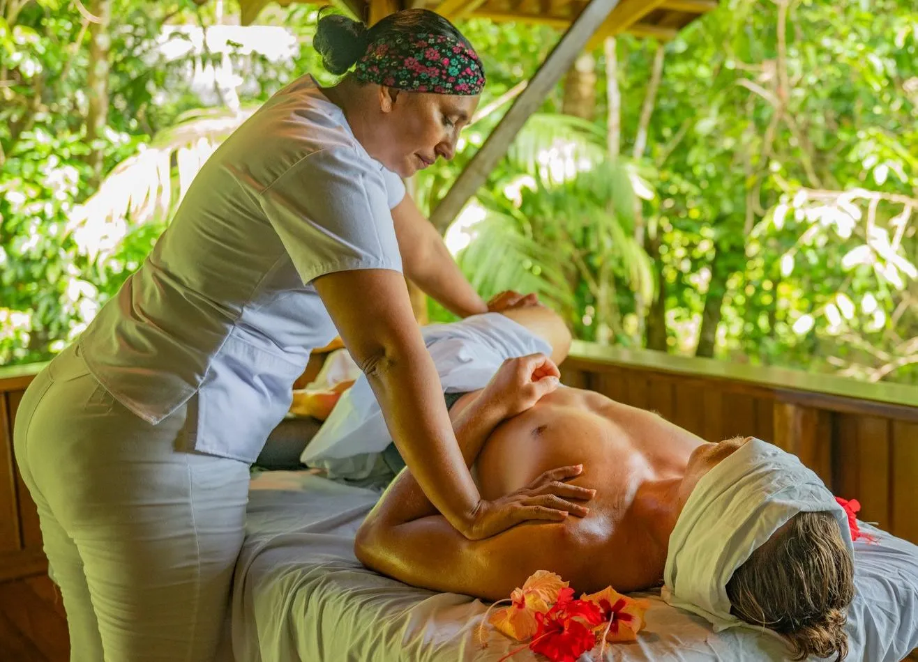 A person receives a massage from a therapist on an outdoor table surrounded by lush greenery; flower petals decorate the table.