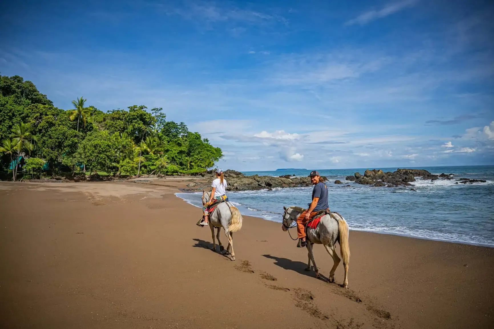 Two people ride horses along a sandy beach near the ocean, with trees and rocks in the background under a clear blue sky.