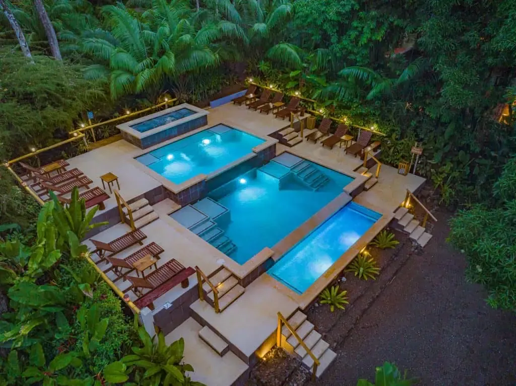 An aerial view of a tiered outdoor swimming pool complex surrounded by tropical plants and trees, with lounge chairs and warm lighting.