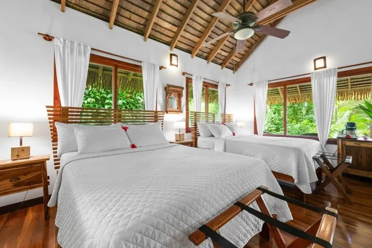 A bright hotel room with two double beds, white bedding, wooden furniture, large windows with white curtains, and a ceiling fan under a thatched roof.