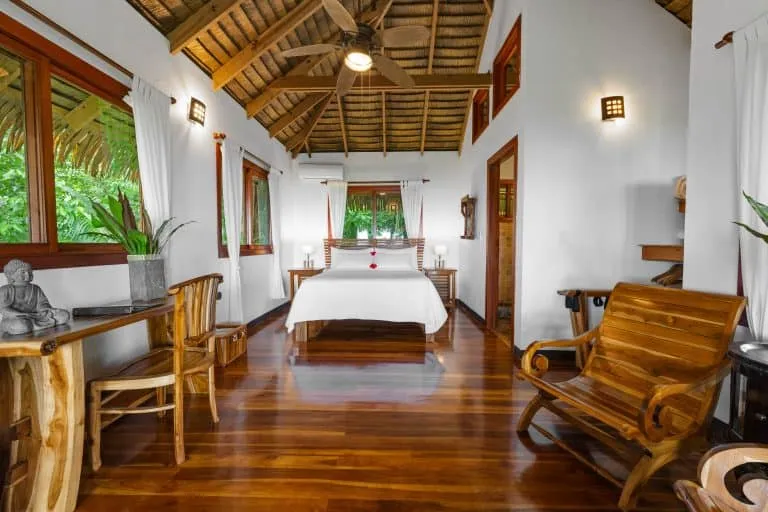 A wooden-floored bedroom with a double bed, wooden furniture, white walls, high ceiling with exposed beams, and large windows overlooking greenery.