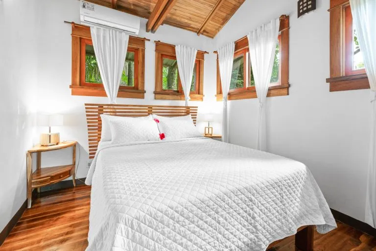 A neatly made bed with a white quilt and pillows, two red flowers on top, wooden floor, white curtains, and four small windows in a bright bedroom.