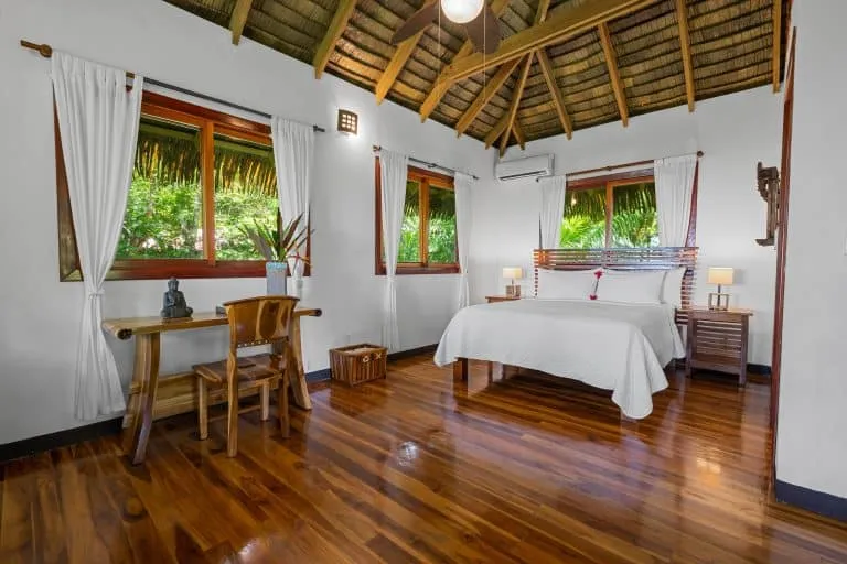 Bedroom with a wooden floor, double bed, bedside tables, desk with chair, large windows with white curtains, and a thatched ceiling.
