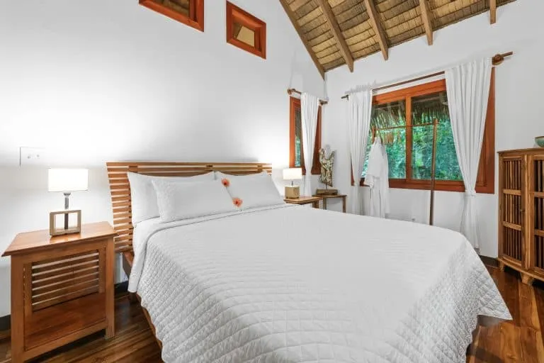 A neatly made bed with a white quilt, flanked by two wooden nightstands with lamps, in a bright room with white walls, wooden floors, and a thatched ceiling.
