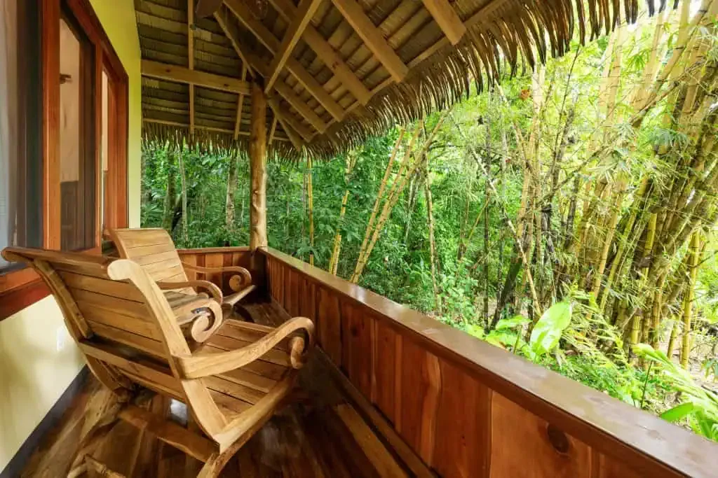 Wooden rocking chairs on a covered balcony with a thatched roof, overlooking dense green bamboo and tropical plants.