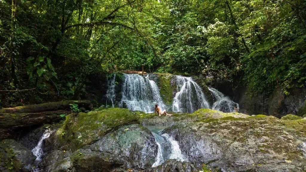 A person sits on rocks at the base of a small waterfall surrounded by dense green forest and moss-covered stones.