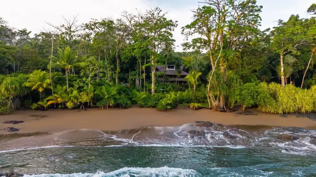 A secluded house surrounded by dense tropical trees sits behind a sandy beach, with gentle ocean waves in the foreground.