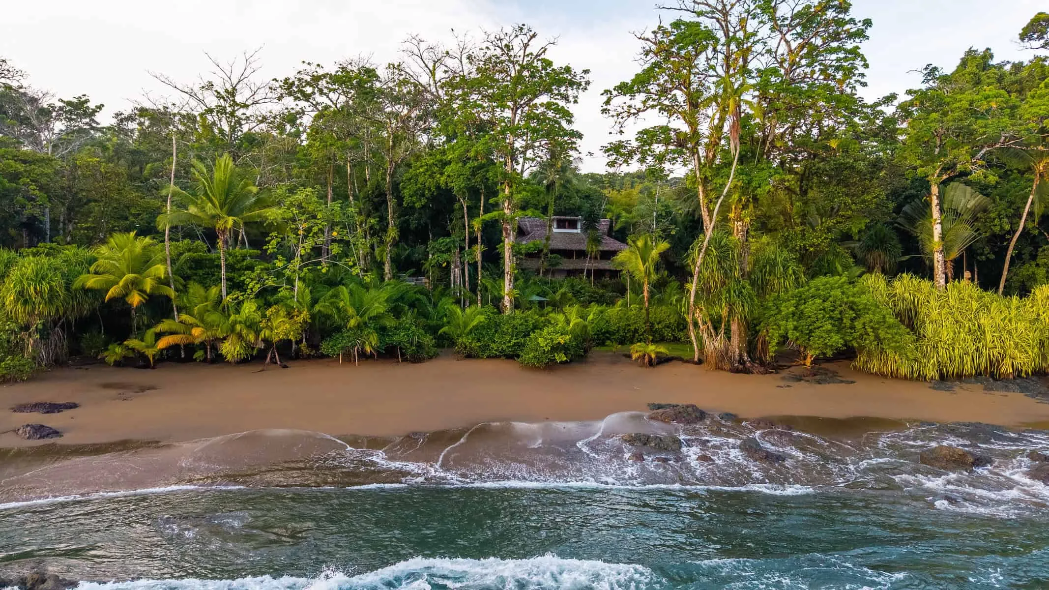 A secluded house surrounded by dense tropical trees sits behind a sandy beach, with gentle ocean waves in the foreground.