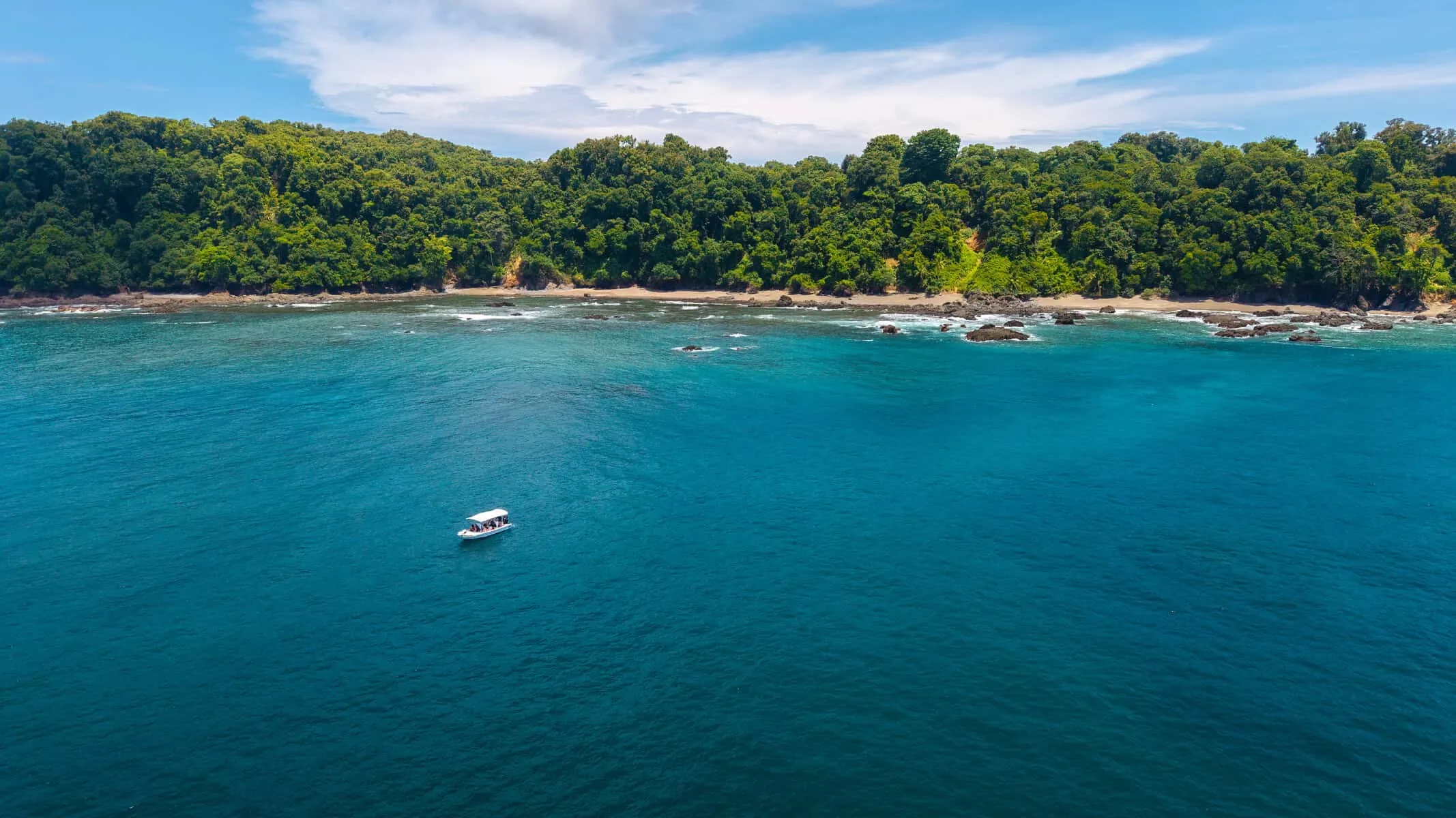 A small white boat floats on clear blue water near a rocky shoreline with dense green trees under a partly cloudy sky.