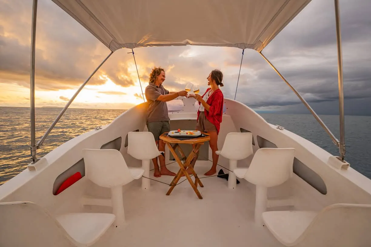 Two people clink glasses on a boat at sunset, standing near a small table with food, surrounded by empty white chairs and calm ocean waters.