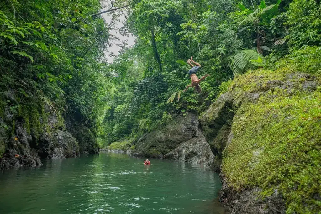 A person swings on a rope and jumps into a river surrounded by lush green forest, while another person swims in the water below.