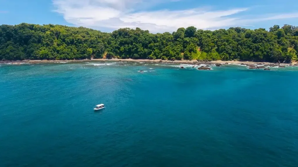 A small boat floats on clear blue water near a forested coastline under a partly cloudy sky.