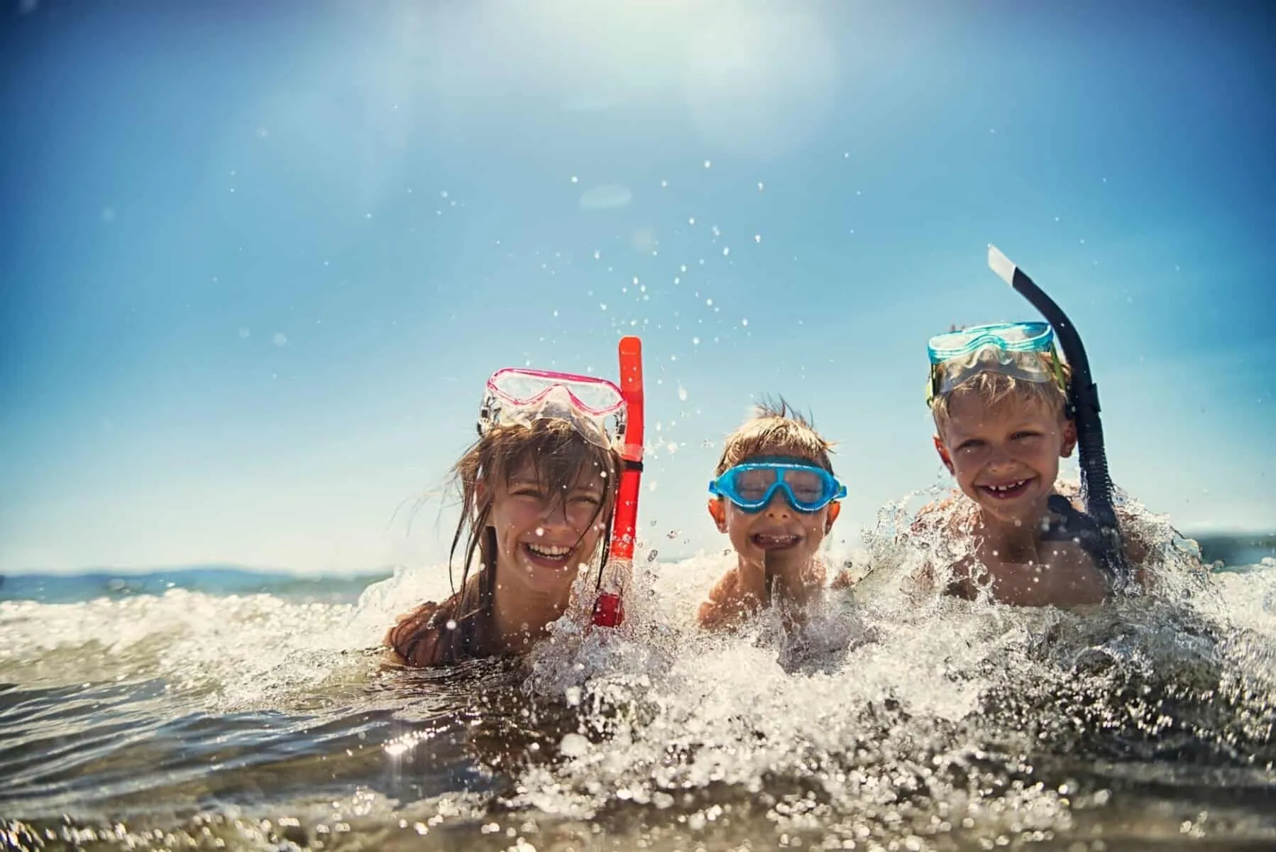 Three children with snorkeling gear splash and play in shallow water under a clear blue sky.