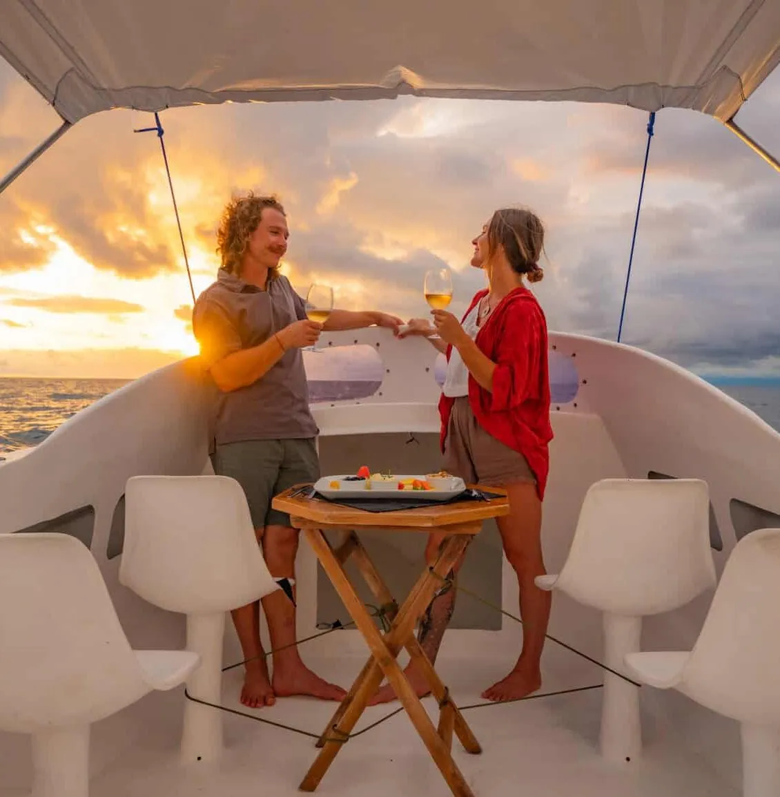 Two people stand on a boat at sunset, clinking glasses and smiling, with a small table of food between them and empty chairs around.
