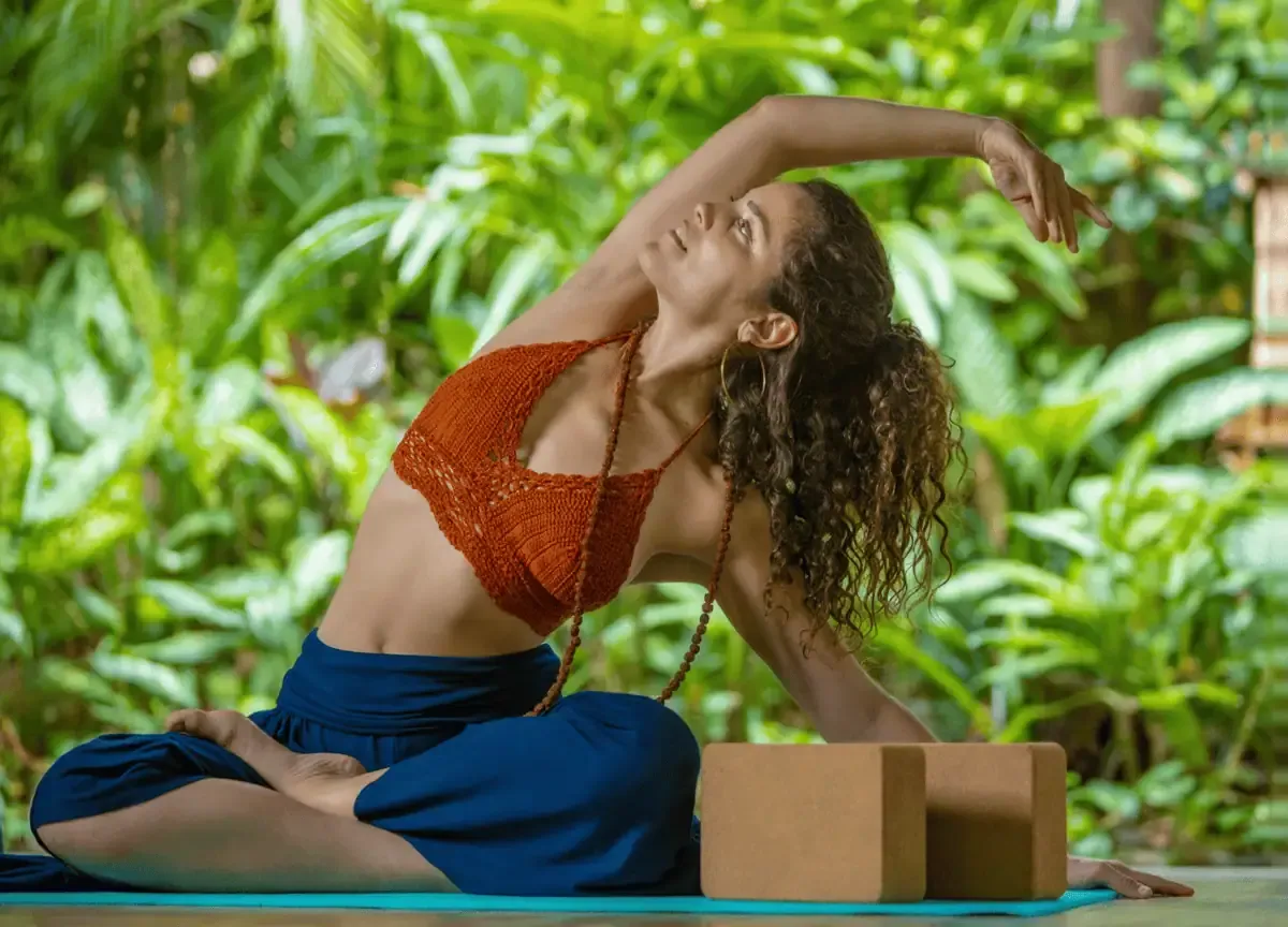 A woman practices a seated side stretch yoga pose on a mat outdoors, surrounded by lush green plants, with two yoga blocks beside her.