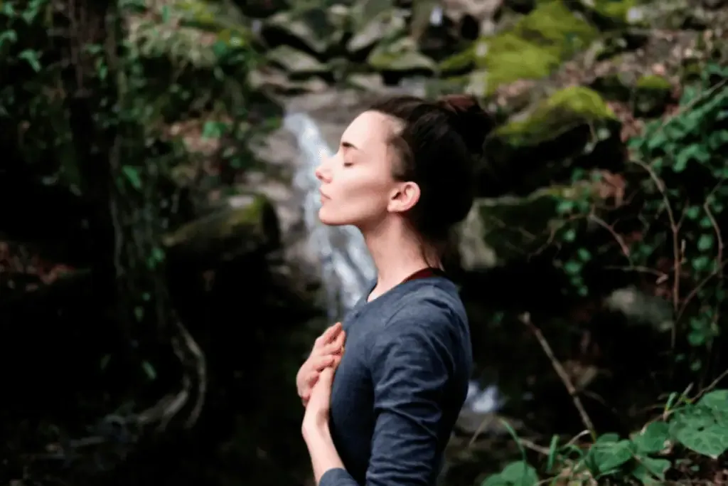 Woman standing outdoors with eyes closed and hands on chest, surrounded by greenery and rocks, appearing to meditate or relax.