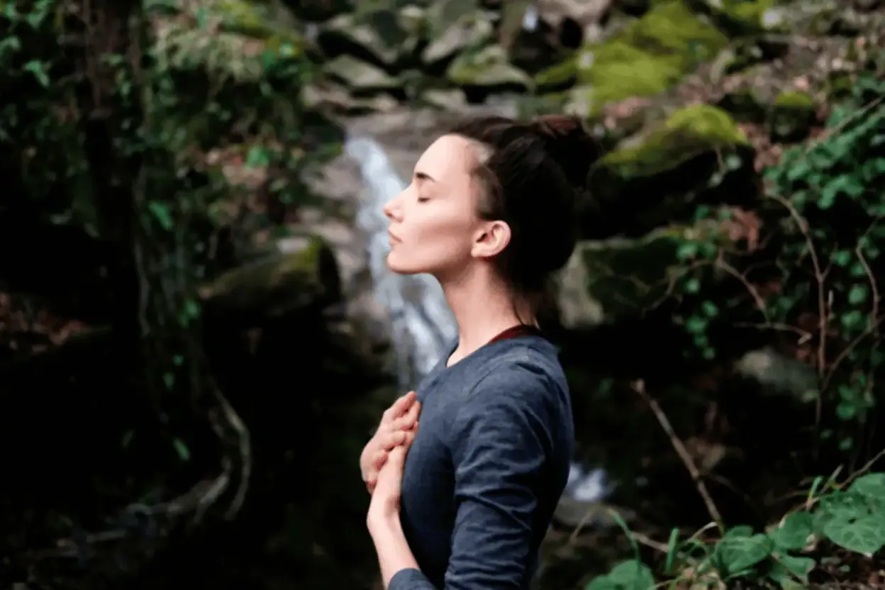 Woman standing outdoors with eyes closed and hands on chest, surrounded by greenery and rocks, appearing to meditate or relax.