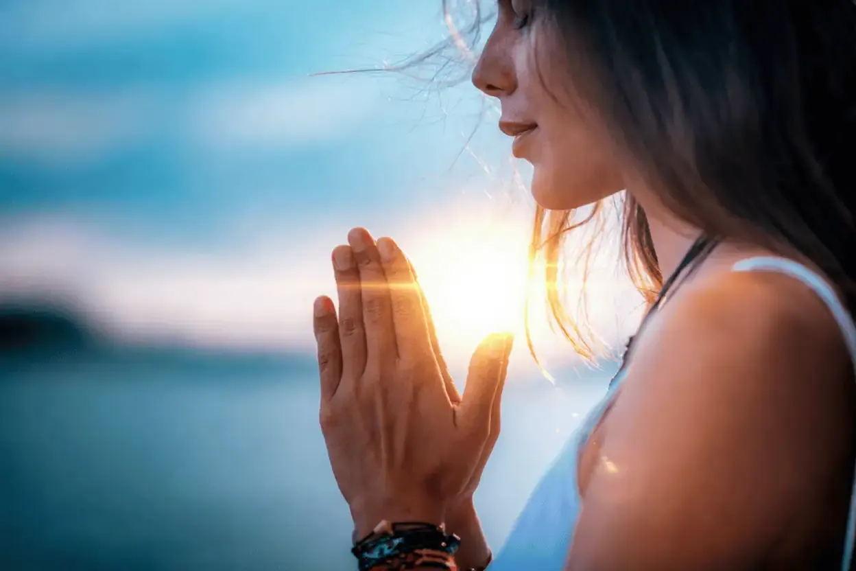 A woman with long hair stands outdoors at sunset, holding her hands together in a prayer position, with sunlight shining between her palms.