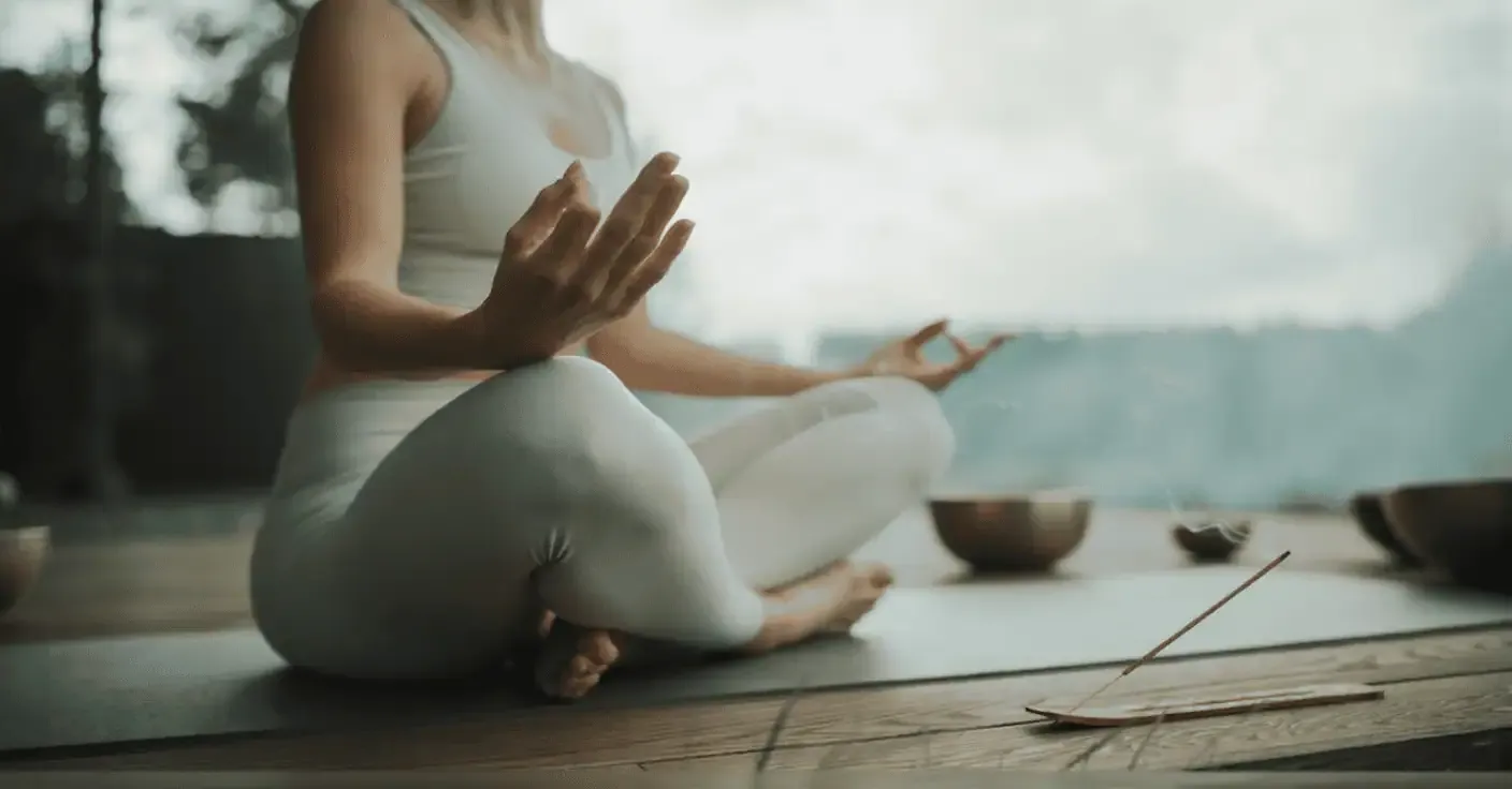A person in white activewear sits cross-legged on a yoga mat, meditating outdoors with incense burning and singing bowls nearby.